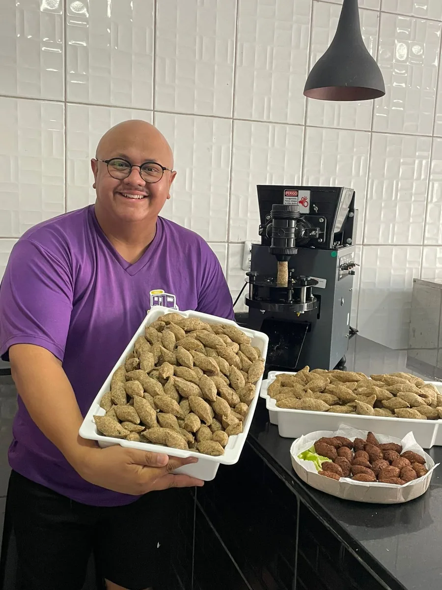 Aluno sorrindo, segurando uma bandeja cheia de quibes modelados, com a modeladora ao fundo e quibes fritos na bancada